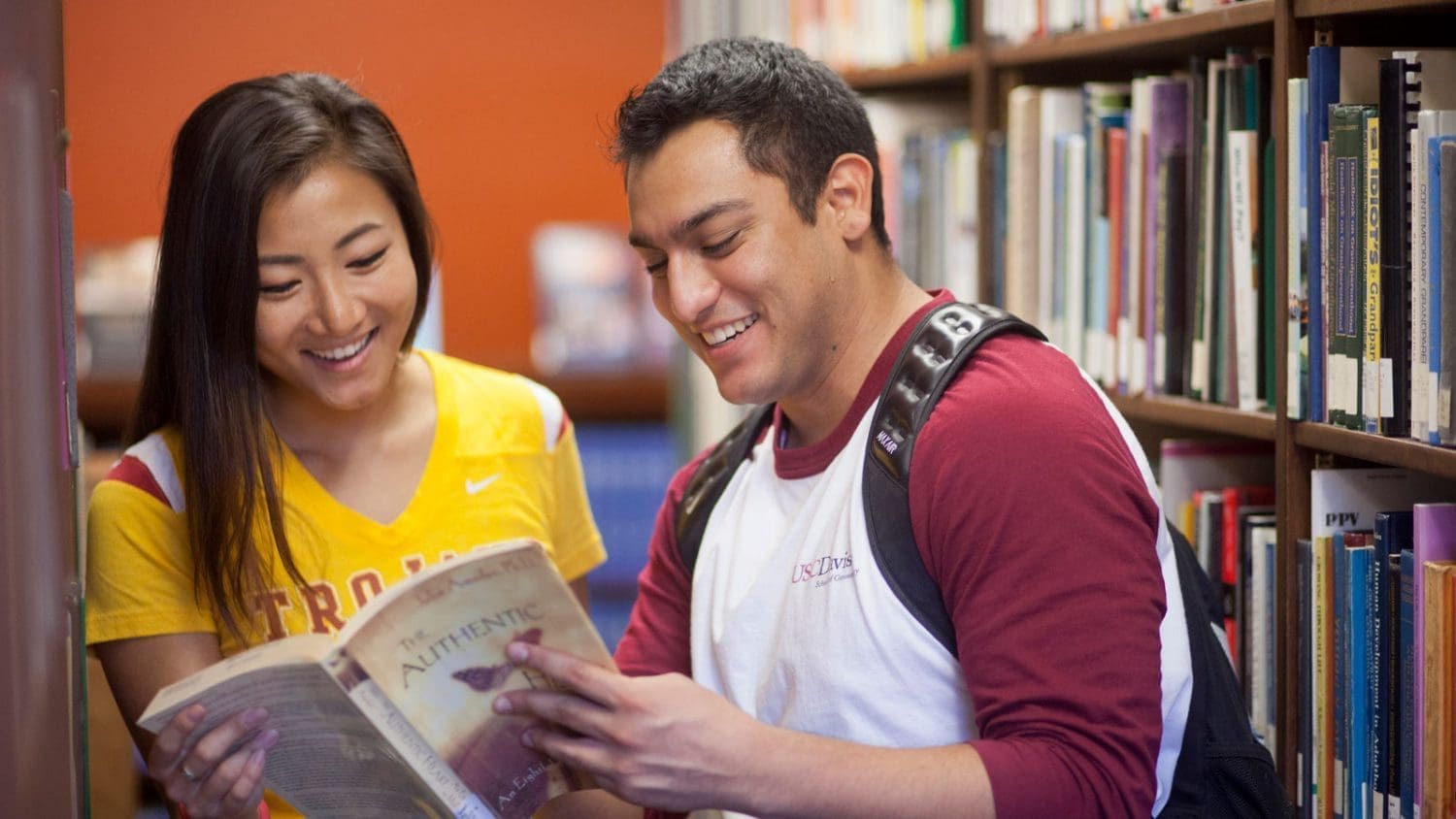 Two students wearing USC shirts, smiling and sharing a book at the library