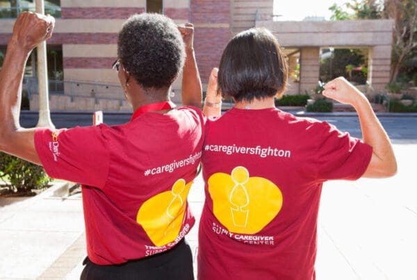 Two women with their backs turned to show their shirts, which say #caregiversfighton and raising their fists