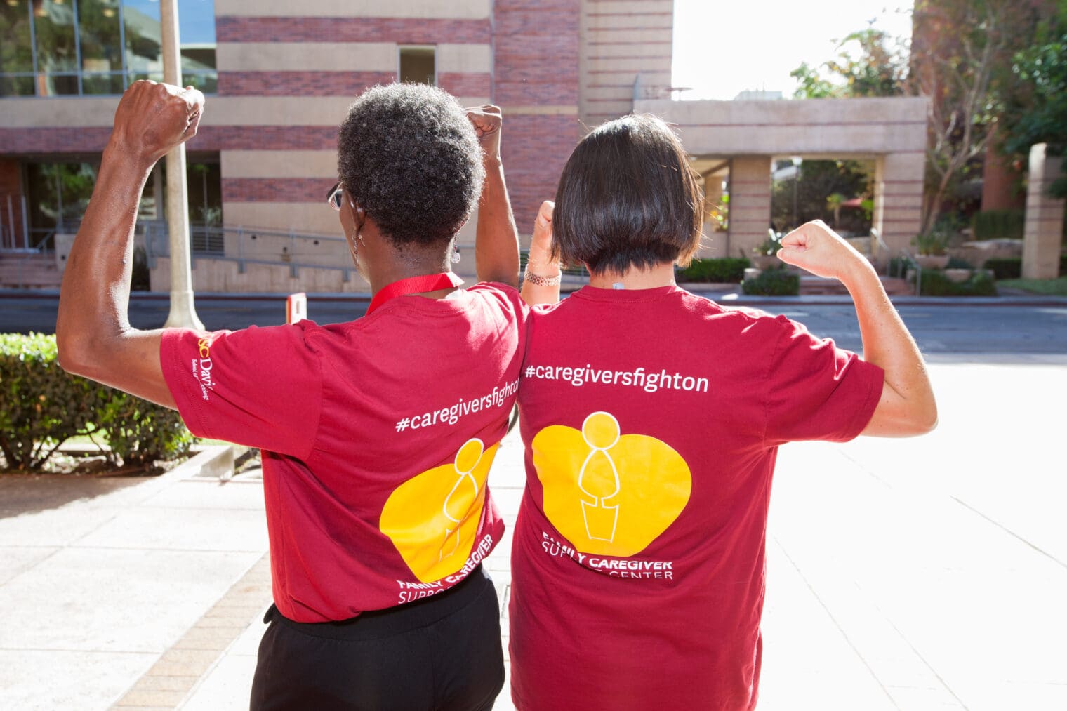 Two women with their backs turned to show their shirts, which say #caregiversfighton and raising their fists