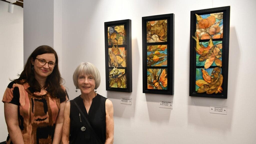 Two women smiling and standing at an art gallery in front of three paintings