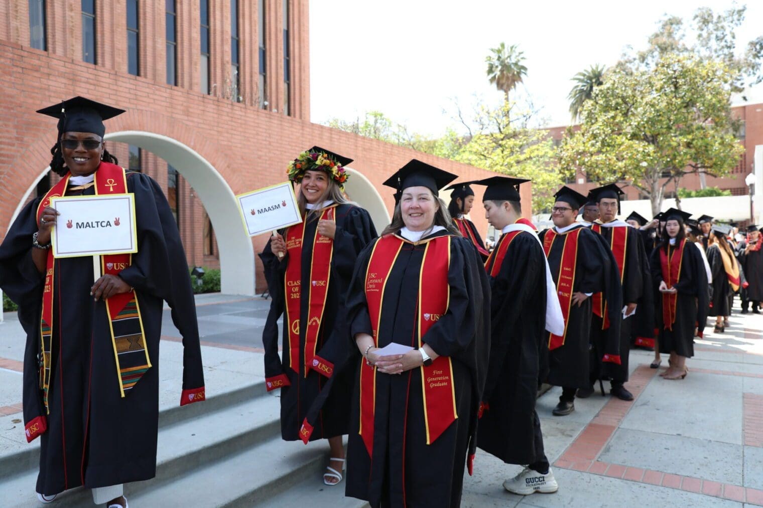 Line of students in commencement robes and caps, with two of them at the front holding signs that depict the degrees they are graduating with. One says "MALTA" and the other says "MASAM."